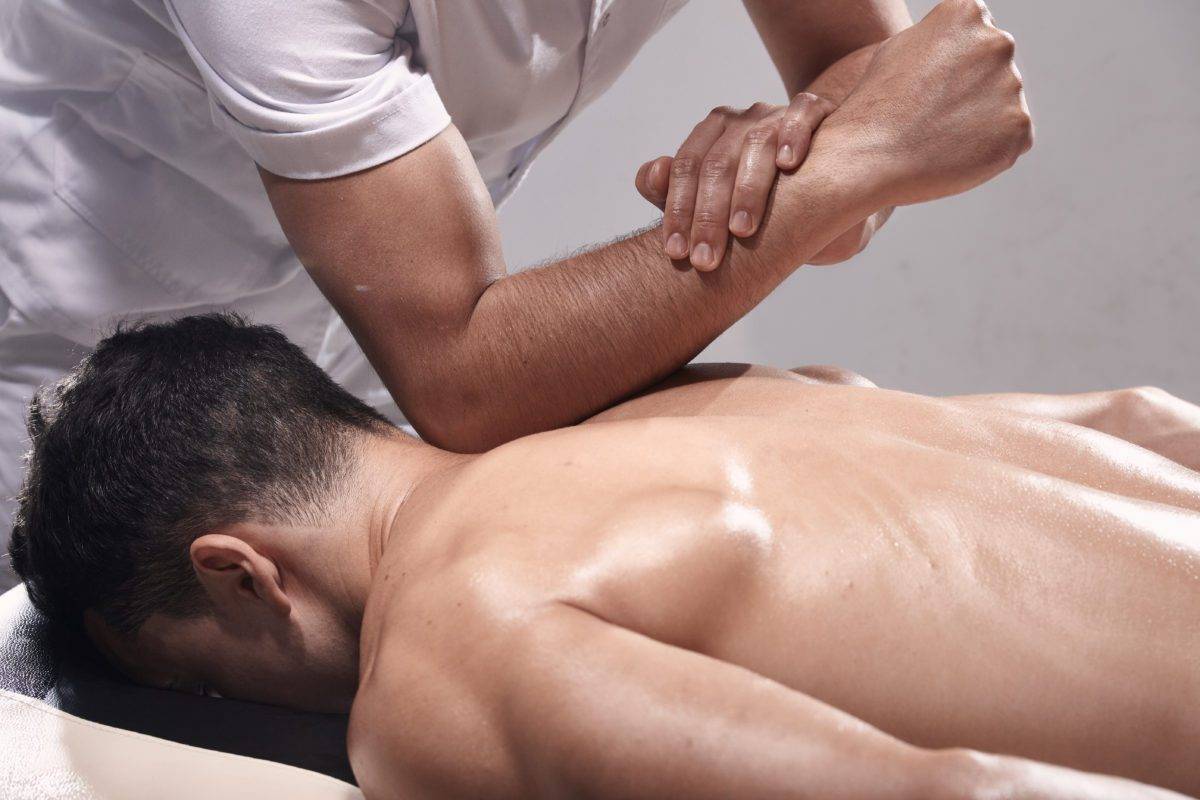 side view, two young man, 20-29 years old, sports physiotherapy indoors in studio, photo shoot. Strong Physiotherapist hard massaging relaxed patient neck back side, with his elbow.