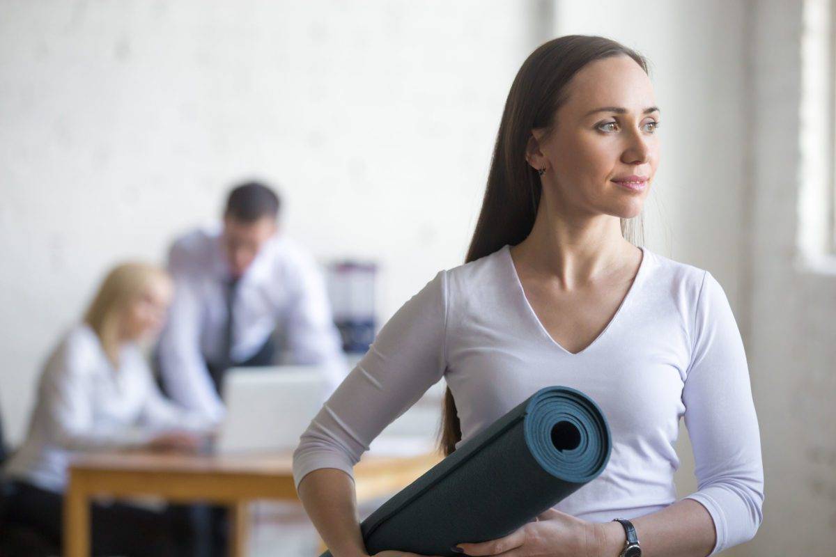 Business and healthy lifestyle concept. Portrait of beautiful sporty young office woman standing with yoga mat at workplace on break time