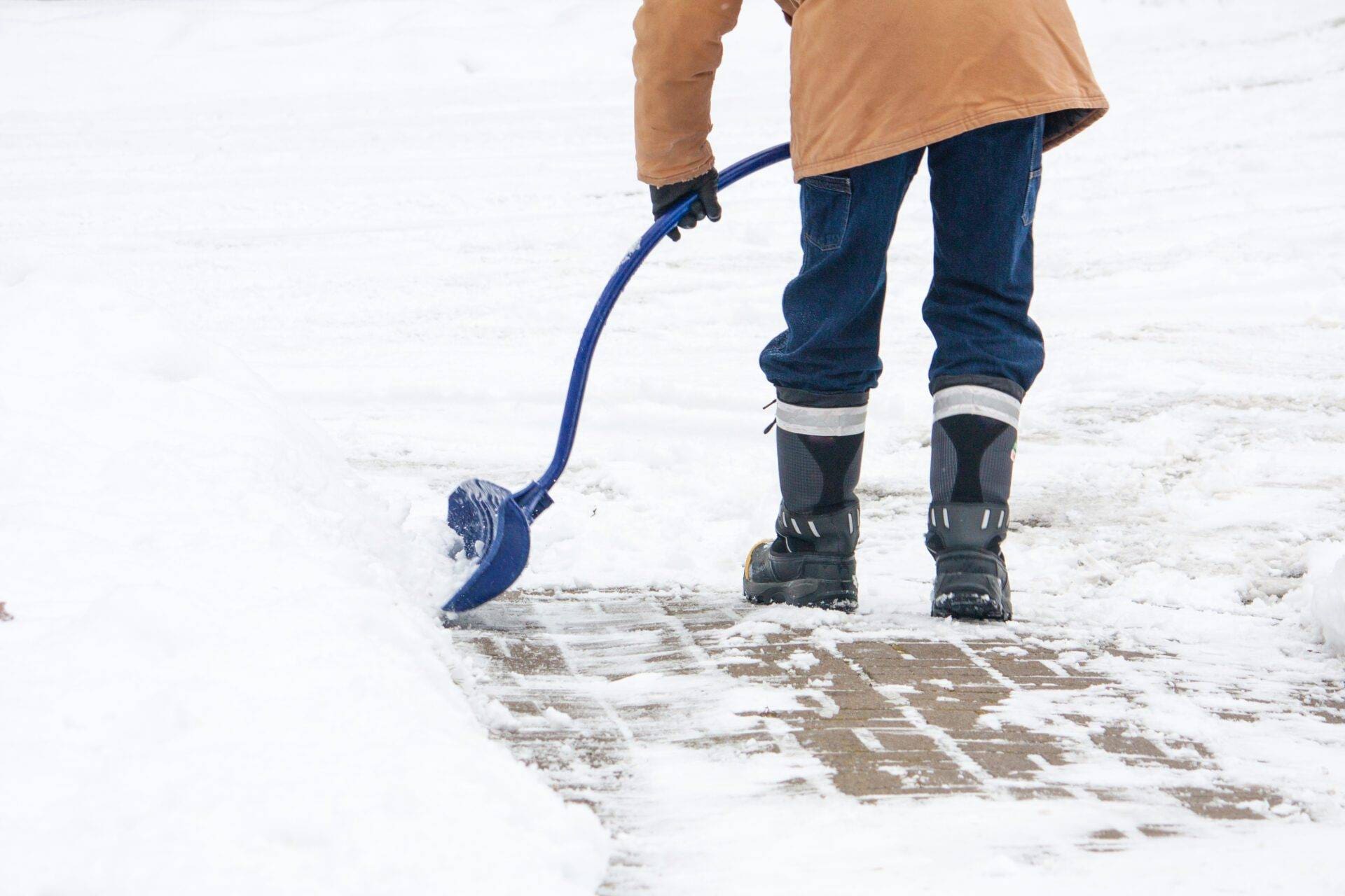 A man with a curved handled snow shovel clearing snow from a brick sidewalk in Canadian winter.